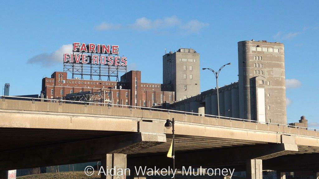 Colour image of a concrete overpass, grain elevator, and a red sign stating "FARINE FIVE ROSES"