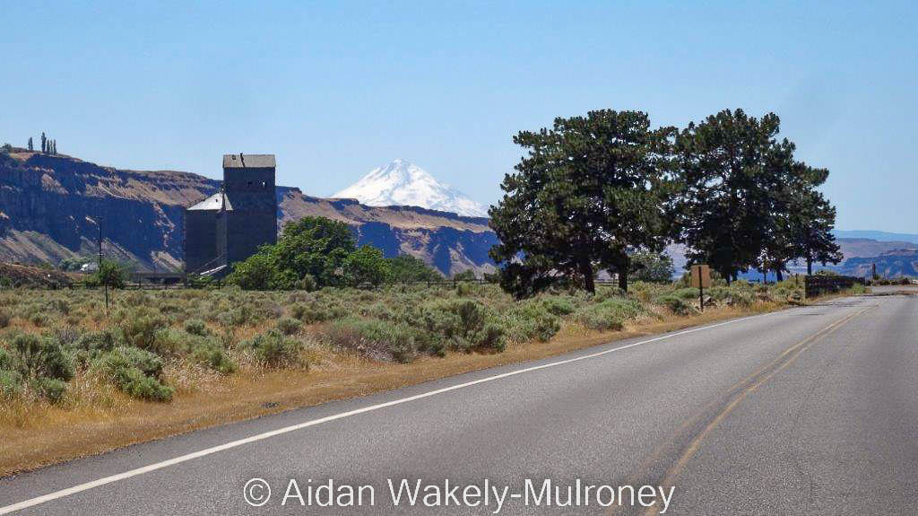 Highway leading to a wooden grain elevator, with a mountain in the background.