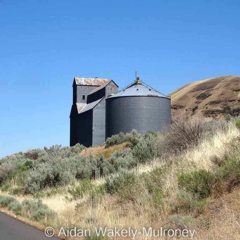 Wooden grain elevator and steel bins on a dry hillside.