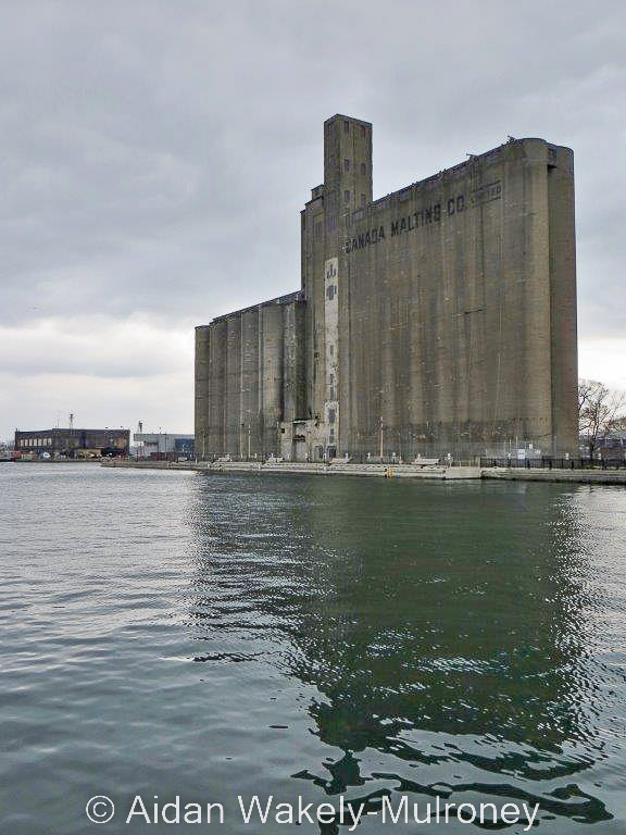 Vertical colour image showing a concrete grain elevator and its reflection on the harbour water. The elevator is lettered CANADA MALTING CO LIMITED.