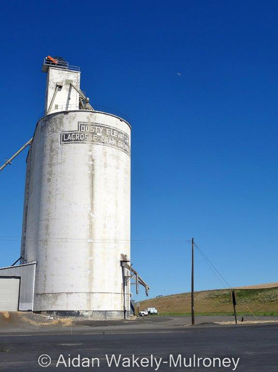 Vertical colour photograph showing a white concrete grain elevator with the words DUSTY ELEVATOR LACROSS GRAIN GROWERS on it.