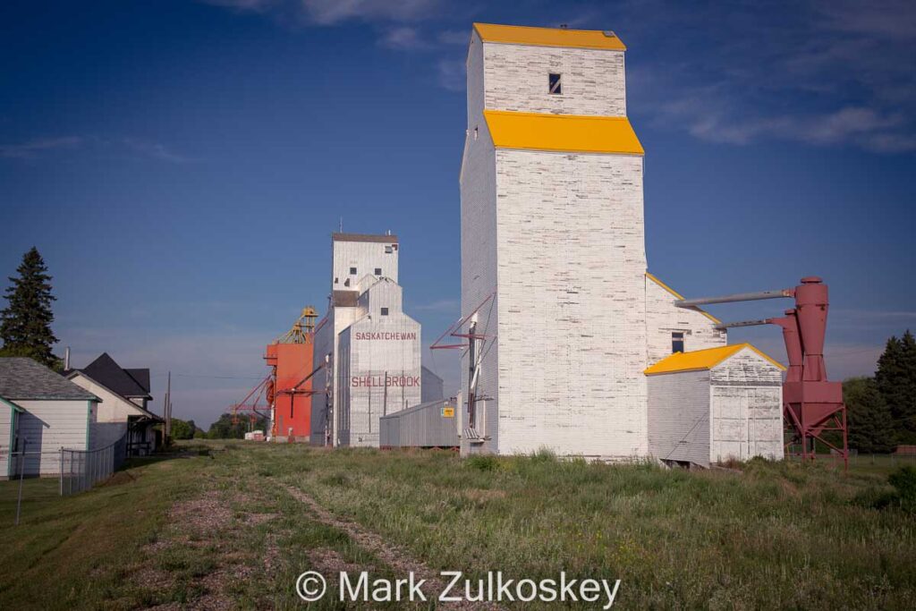 Colour photograph of three wooden grain elevators in a row with a preserved train station across from them.