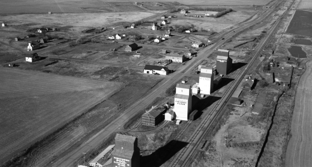 Aerial black and white photograph showing a small town in Saskatchewan with a rail line with four grain elevators and a station.