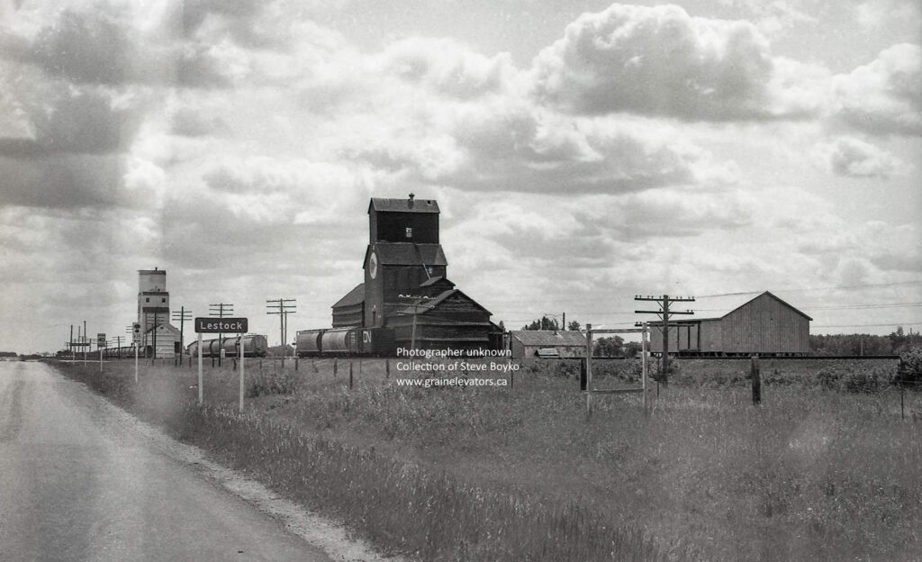 Black and white photograph of two grain elevators and a highway sign "Lestock"