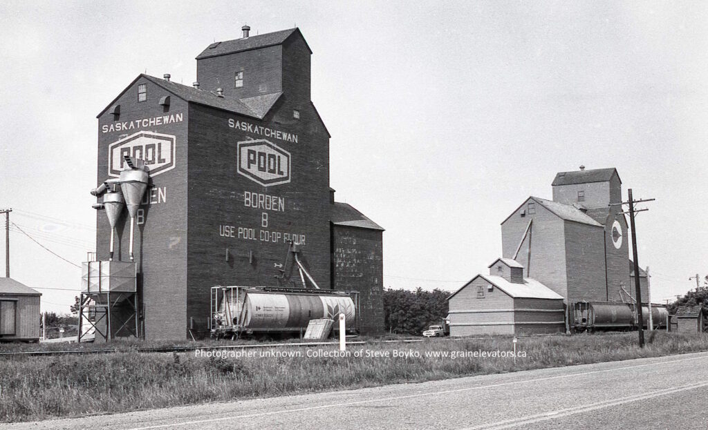 Black and white photograph of two wooden grain elevators with "Saskatchewan Pool" on it and rail cars in front
