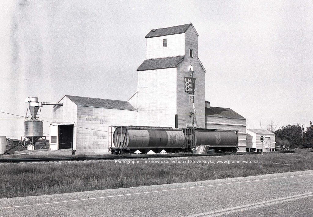 White wooden grain elevator with "United Grain Growers" on it and rail cars in front
